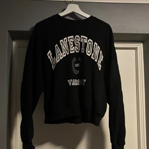 Svart sweatshirt från NLY Trend - Säljer en svart sweatshirt från NLY Trend med trycket 'LANESTONE VARSITY' i vitt. Tröjan har en klassisk rund hals och långa ärmar, perfekt för en avslappnad stil. Den är i bra skick och passar perfekt för höst och vinter.