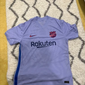 Lila FC Barcelona fotbollströja - Säljer en lila FC Barcelona fotbollströja från Nike i mycket bra skick. Tröjan är kortärmad med klubbens emblem och sponsorer tryckta på framsidan. Perfekt för fans som vill visa sitt stöd på matchdagar! 💜