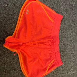 Träningsshorts under armour - Snygga korta träningsshorts i korall/orange storlek S. Från Under armour. Shortsen har en resår i midjan och en dragsko för att justera passformen. De har även fickor på sidorna. Några små tråddragningar och fläckar som jag tror går bort i tvätten. 