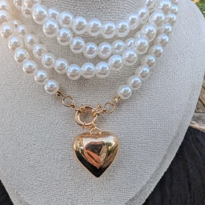 Vackert pärlhalsband med hjärthänge - Elegant pärlhalsband med flera rader av vita pärlor och ett stort guldfärgat hjärthänge. Perfekt för att ge en klassisk touch till vilken outfit som helst.