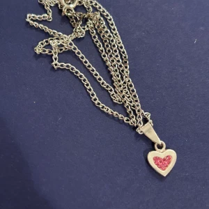 Silverhalsband med hjärthänge - Säljer ett superfint silverhalsband med ett hjärthänge. Hänget har en söt rosa glittrig detalj i mitten som verkligen poppar! Perfekt för att ge en romantisk touch till vilken outfit som helst. Kedjan är tunn och elegant, vilket gör den lätt att bära varje dag.