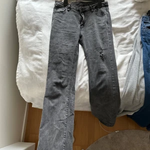 Grå jeans med slitningar - Säljer ett par grå lågmidjade bootcut jeans med coola slitningar på ena benet. De har en klassisk femficksdesign och en dragkedja med knapp i midjan. Perfekta för en avslappnad och trendig look! Passar mig som är 175!
