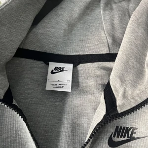 Grå hoodie från Nike - Säljer denna grå nike tech hoodien i storlek S. Den har en dragkedja framtill och en klassisk Nike-logga på bröstet. Perfekt för både träning och vardag. Hoodien är långärmad och har en bekväm passform. Använd men bra skick, pris kan diskuteras!