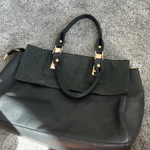 Svart handväska med gulddetaljer - Säljer en elegant svart handväska med stilrena gulddetaljer. Väskan har två robusta handtag och en praktisk dragkedja. Perfekt för både vardag och fest!