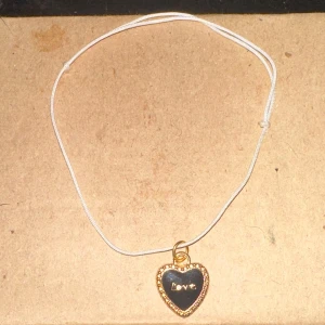 Armband med hjärthänge - Säljer ett sött armband med ett hjärthänge. Hänget är svart med en guldig kant och har texten 'love' ingraverad. Bandet är enkel och vit, vilket gör det perfekt för vardagsbruk eller som en fin present.