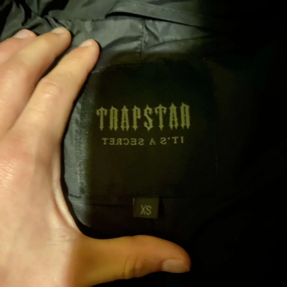 Säljer en ny svart pufferjacka från Trapstar i storlek XS. Jackan har en cool broderad text på ryggen och en dragkedja framtill. Den är perfekt för vintern med sin varma och quiltade design. Huvan ger extra skydd mot kylan. Tar även emot prisförslag!. Jackor.