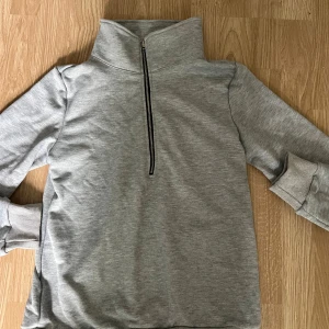 Grå tröja med dragkedja - Säljer en stilren grå tröja med halv dragkedja framtill. Den har långa ärmar och ribbade muddar vid ärmslut och nederkant. Perfekt för en avslappnad look under höst och vår.
