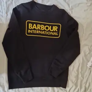 Säljer en snygg svart tröja från Barbour International. Den har ett coolt gult tryck på framsidan med märkesnamnet. Tröjan är i storlek M och är gjord av 85% bomull och 15% polyester, vilket gör den både bekväm och hållbar. Perfekt för en casual look!