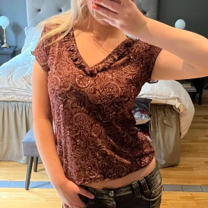 Brun mönstrad topp - Säljer en superfin vinröd topp med ett snyggt paisleymönster. Den har korta ärmar och en v-ringad hals med volangdetaljer. Perfekt för både vardag och fest! Står tyvärr inte märke eller storlek. Skulle gissa på S eller M.