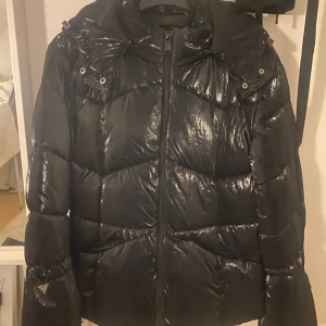 Svart pufferjacka från Guess - Säljer en snygg svart pufferjacka från Guess. Den är perfekt för kalla dagar med sitt blanka material och quiltade design. Jackan har en dragkedja framtill och en mysig huva. Den är i bra skick och redo för vintern!