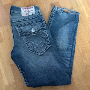 Säljer ett par snygga blå jeans från True Religion. De har en cool slitning och klassiska bakfickor med knappar och broderad detalj. Perfekta för en casual look!