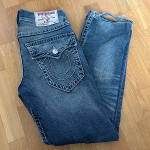 Blå jeans från True Religion - Säljer ett par snygga blå jeans från True Religion. De har en cool slitning och klassiska bakfickor med knappar och broderad detalj. Perfekta för en casual look!