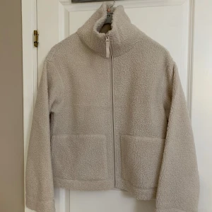 Beige teddyjacka med dragkedja - Säljer en supermysig beige teddyjacka som är perfekt för kyliga dagar. Jackan har en dragkedja framtill och stora fickor för att hålla händerna varma. Den är långärmad och har en hög krage för extra värme. Varan är oanvänd med etiketten kvar.