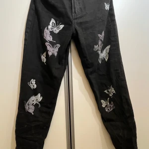 Svarta jeans med fjärilsmönster - Säljer ett par svarta högmidjade mom jeans med snygga vitaoch lila fjärilar tryckta längs benen. Använda fåtal gånger, men lite av trycket har blivit slitet i tvätt. 