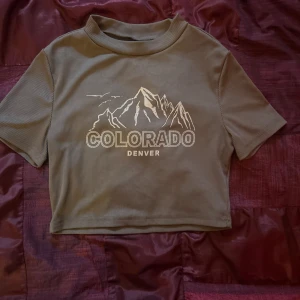 Grön croppad topp med Colorado-tryck - Säljer en  beige croppad topp med tryck av berg och texten 'Colorado Denver'. Toppen har korta ärmar och en ribbad struktur. Perfekt för en avslappnad stil eller en dag ute i solen!