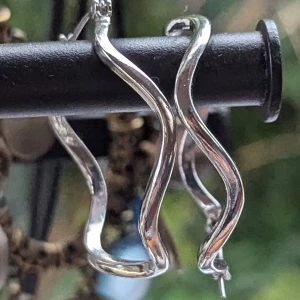 Silvriga örhängen i oregelbunden form - Örhängen med en unik, oregelbunden form. Örhängena är runda men har en vågig design som ger dem en modern och stilren look. De är tillverkade i ett glänsande silvermaterial och har en enkel låsmekanism.