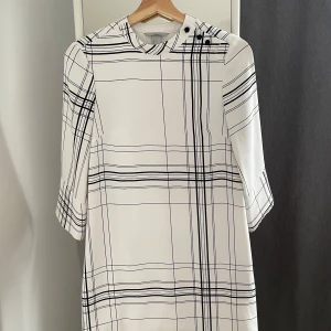 Vit och svart rutig klänning från H&M - Säljer en superfin vit klänning med svart rutmönster från H&M. Den har trekvartsärmar och en stilren design med knappar vid axeln. Perfekt för både vardag och festliga tillfällen. Klänningen är i storlek XS och sitter bekvämt med en normal passform.