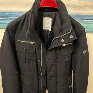 Svart jacka från J.Lindeberg - Säljer en snygg svart jacka från J.Lindeberg i storlek M. Jackan har en stilren design med dragkedja och knappar framtill. Den har flera praktiska fickor med dragkedjor och knappar, perfekt för höst och vinter. Jackan är i bra skick och redo för nya äventyr!