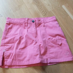 Rosa kortkjol - Säljer en superfin rosa kortkjol med en liten slits och broderad logga. Perfekt för sommardagar! Kjolen har praktiska fickor och en knapp i midjan, den har även inbyggda shorts i kjolen! Den är i bra skick och redo för nya äventyr. Passar perfekt till en avslappnad stil eller en dag på stan. 🌸 Den är i storlek 150 (men passar mig som har 170) och är köpt på Pelle P Hör av er om frågor!💕