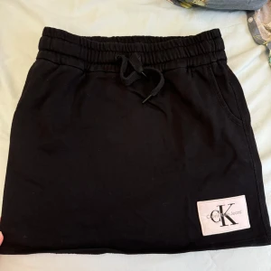 Svarta kjol från Calvin Klein Jeans - Säljer min snygga svarta kjol från Calvin Klein Jeans i storlek XS. De har en bekväm elastisk midja med knytband och en stilren design med en logga på benet. Perfekta för en avslappnad sommarlook!