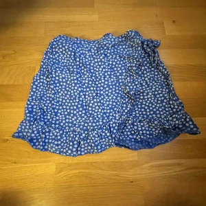 Blå blommig kjol med volang - Säljer en superfin blå kjol med vita blommor. Den har en söt volangdetalj längst ner som ger den en extra touch. Perfekt för sommardagar eller en mysig vårutflykt. Kjolen är i bra skick, köpt på marknad i Spanien så har ingen storlek