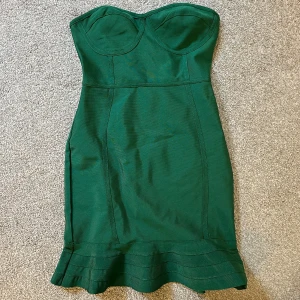 Grön axelbandslös klänning - Snygg grön axelbandslös bandage klänning i tight passform. Perfekt för festliga tillfällen med sin eleganta design och volangdetalj vid fållen. Klänningen är i nyskick och har en smickrande siluett.