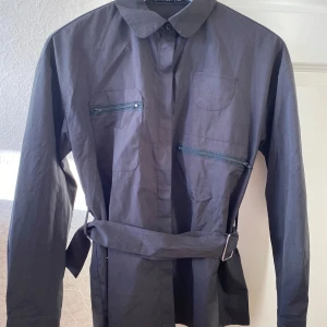 Svart jacka med bälte - Säljer en stilren svart skjorta med bälte i midjan. Skjortan har långa ärmar och två dragkedjefickor på bröstet. Perfekt för höst och vår!