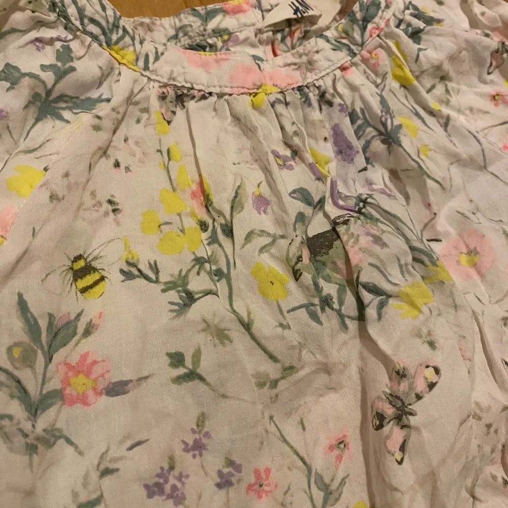 Säljer en söt blommig topp från H&M i storlek 122. Toppen är vit med ett färgglatt blommönster i rosa, gult och grönt, samt små bin och fjärilar. Den har en lös passform och är ärmlös, perfekt för varma sommardagar. Bra skick och redo för ny ägare!. Toppar.