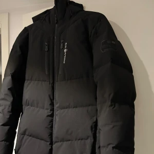 Svart dunjacka från Sail Racing - Säljer en svart dunjacka från Sail Racing i storlek medium. Jackan har en dragkedja framtill och är perfekt för kalla vinterdagar. Den är i mycket bra skick och har en stilren design med en logga på bröstet. 