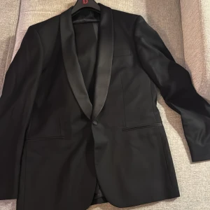  Smoking kostym från Dressmann - Elegant svart kavaj med matchande byxor från Dressmann. Kavajen har en klassisk design med satinslag och en knapp framtill. Byxorna har en stilren look med knappstängning. Perfekt för formella tillfällen.  Byxornas storlek är 31-32, kavajen är 48 och skjortan M