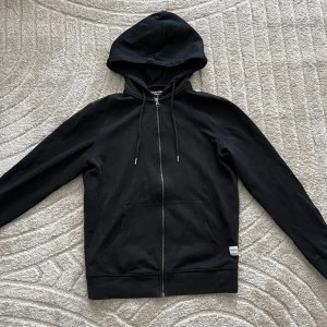 Svart hoodie med dragkedja - Säljer en stilren svart hoodie med dragkedja och justerbar huva. Tröjan har långa ärmar och praktiska fickor framtill. Perfekt för en avslappnad look.