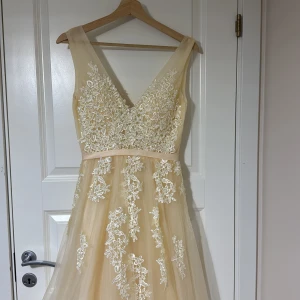 Champagnefärgad långklänning med spetsdetaljer - Elegant champagnefärgad långklänning med v-ringning och vackra spetsdetaljer. Klänningen har en ärmlös design och ett skirt lager som ger en drömlik känsla. Perfekt för speciella tillfällen.