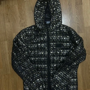 EA7 Emporio Armani Jacka - Svart dunjacka från Emporio Armani med guldigt EA7-mönster över hela jackan. Använt Max 3 gånger. Nypris: 1900kr 
