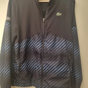 Svart och blå randig jacka från Lacoste - Svart och blå randig jacka från Lacoste Sport. Jackan har dragkedja och långa ärmar med ett Lacoste-märke på bröstet. Två små hål vid vänster ficka men finns ett lager till bakom som är svart så de syns knappt.