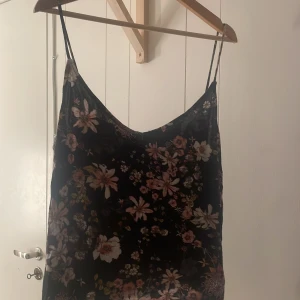 Blommig linne från H&M - Säljer ett svart linne från H&M med vackert blommönster i rosa och beige. Linnet har tunna axelband och en luftig passform, perfekt för varma dagar.