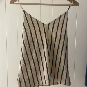 Randig linnetopp från Gina Tricot - Snygg randig linnetopp från Gina Tricot i beige och svart. Toppen har smala axelband och ett korsat band i ryggen för en stilren look. Perfekt för varma sommardagar.