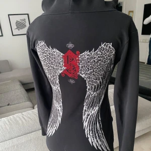 Svart hoodie med vingar från Sinful - Snygg svart hoodie från Sinful med unikt tryck av vita vingar och röd detalj på ryggen. Tröjan har en djup v-ringning och en praktisk magficka framtill. Perfekt för en cool och avslappnad stil. Och rhinestones på hela ryggen  perfekt skick alla rhinestones är kvar