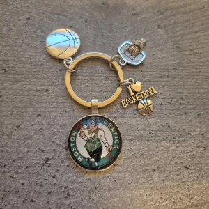 Nyckelring med Boston Celtics tema - Cool nyckelring med Boston Celtics tema. Den har flera hängen, inklusive en basketboll, en basketkorg och en medaljong med Celtics-loggan. Perfekt för basketfans!