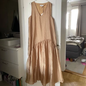 Champagnefärgad långklänning från Joelle - Elegant champagnefärgad långklänning från Joelle med v-ringning och ärmlös design. Köpt på Ellos för 799kr. Tillverkad i ett glittrande material som ger en lyxig känsla. Perfekt för speciella tillfällen. Storlek 38/40. Helt NY med lappar! 