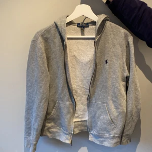 Grå hoodie från Ralph Lauren - Säljer en klassisk grå hoodie från Ralph Lauren med dragkedja och huva. Tröjan har långa ärmar och en liten broderad logotyp på bröstet. Perfekt för en avslappnad stil.
