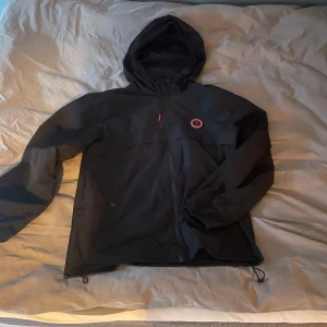 Svart jacka från Trapstar - Snygg svart jacka från Trapstar med huva och dragkedja. Jackan har ett coolt tryck på ryggen och en logotyp på bröstet. Perfekt för en stilren look.