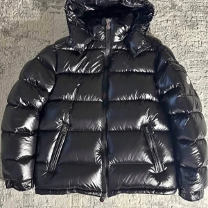 Svart moncler maya  - Snygg svart pufferjacka med glansig yta och praktisk huva. Jackan har dragkedja framtill och två fickor med dragkedjor. Perfekt för kyliga dagar.