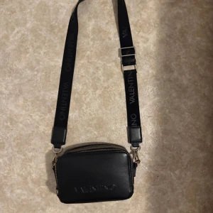 Svart axelväska från Valentino - Nu säljer jag min snygga svarta axelväska från Valentino med justerbar rem och dubbla dragkedjor. Perfekt för att bära dina nödvändigheter med stil. Väskan har en elegant design med märkets logga på remmen, och i framsidan. Köpt hos Zalando för ungefär 1200kr säljer för 700kr. De ända ”defekterna” är att den har rostat lite i dragkedjan. Det går enkelt att fixa med lite färg! Finns fler bilder vid önskning. Den är ganska stor och rymmer mycket. Pris går att diskutera! 