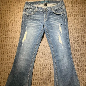 Blå flare jeans från Ariya - Snygga blå flare jeans från Ariya med slitna detaljer på framsidan. Och dekorativa detaljer på baksidan, de är flare. Midja rakt över: 41cm. Lår: 27cm. Innerben: 81