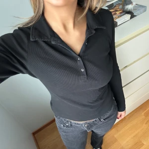 Svart ribbad pikétröja - Snygg svart ribbad pikétröja med knappar framtill. Perfekt för en stilren look. Tröjan har långa ärmar och en klassisk krage.