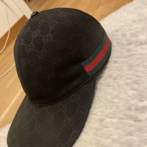 Svart keps med mönster och ränder - Snygg svart keps från Gucci med diskret mönster och ikoniska gröna och röda ränder på sidan. Justerbar rem baktill för perfekt passform. Perfekt för en stilren look.