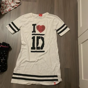 Vit nattklänning med tryck 1D - Säljer en vit nattklänning med svart och rött tryck 'I ❤️ 1D'. Den har korta ärmar med svarta ränder och en avslappnad passform. Hjärtat är slitet för glittret hade åkt av
