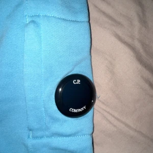 Blå hoodie från C.P. Company - Säljer en snygg blå hoodie från C.P. Company. Den har en klassisk design med en ficka framtill och en huva. Detaljerad med en rund logotyp på ärmen. Perfekt för en avslappnad stil. Köpt för 2000 men säljs för 600 nästan helt ny. Äkta