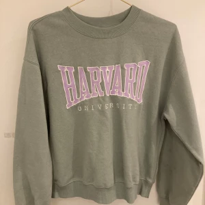 Grön sweatshirt med Harvard-tryck från Divided - Säljer en grön sweatshirt från Divided med ett stort lila Harvard University-tryck på framsidan. Tröjan har en rund halsringning och långa ärmar. Perfekt för en avslappnad stil!