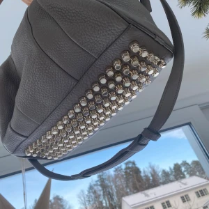 Grå axelväska från Alexander Wang med nitar - Snygg grå axelväska i skinn från Alexander Wang. Väskan har coola nitar på undersidan och en praktisk dragkedja. Perfekt för att ge din outfit en edgy touch. Väldigt bra skick. Äkta men har inget kvitto då jag haft den ett tag. Kan skicka fler bilder vid intresse! Modell: Peddled Lambskin Rocco Oyster 